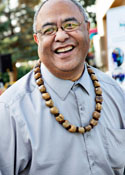 A Polynesian man wearing a blue button down and a wooden necklace smiles at the camera