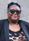 A black woman wearing sunglasses and a black blazer stands in front of a grey wall