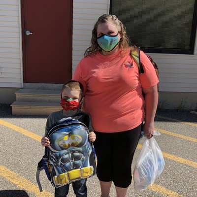 A woman stands with a child holding a backpack. Both are wearing facemasks.