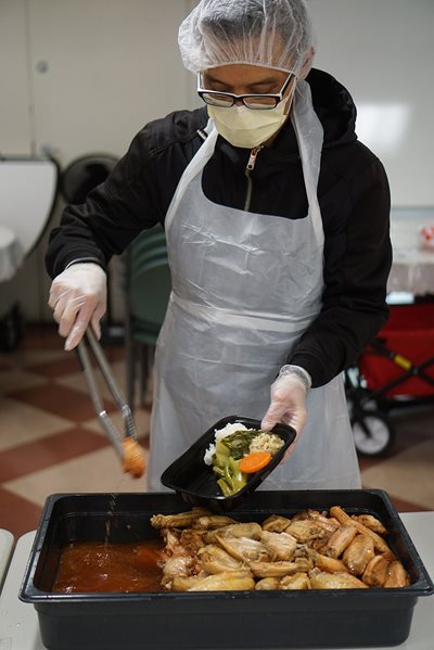 A worker, in a mask and hairnet, is ready to distribute food.