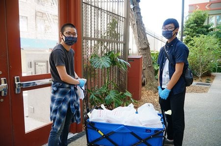 Making deliveries in the community for Chinatown CDC, two staff members stand near a big blue cart filled with food items.