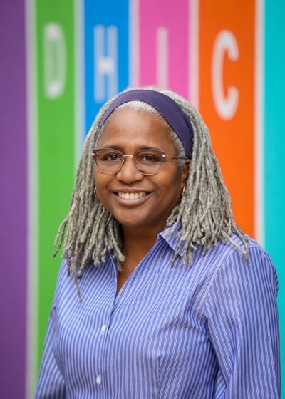 Yolanda Winstead stands against a background that has a rainbow of colors. She has long hair, braided, and glasses and a broad smile.