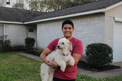 Alex holds his dog and smiles at the camera in front of his home.