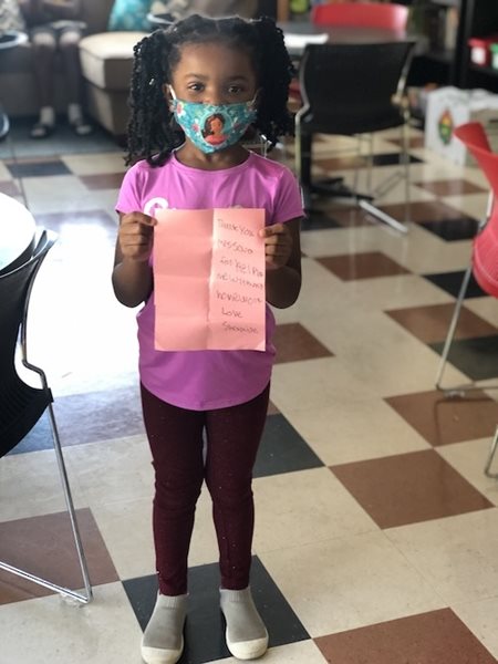 A young girl in a mask holds up a sign, thanking staff members.