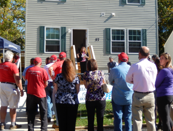 A crowd of people standing outside of a home and listening to a speech.