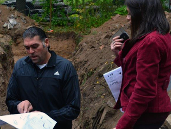 A man wearing a black jacket points at a map. A woman wearing a red jacket holds her cell phone and another map.