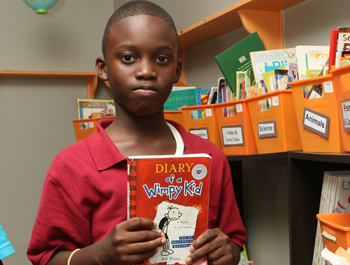 A black boy wearing a red polo shirt holds up a book titled "Diary of a Wimpy Kid"