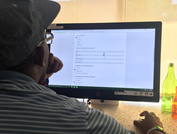 A black man wearing a black baseball cap and a striped shirt sits in front of a computer