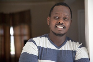 Alain Rwage, a black man, stands in his home wearing a blue and white striped shirt